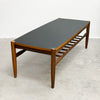 A mid-century modern wooden coffee table with a dark gray rectangular top and a lower slatted shelf, set against a plain white wall and concrete floor.