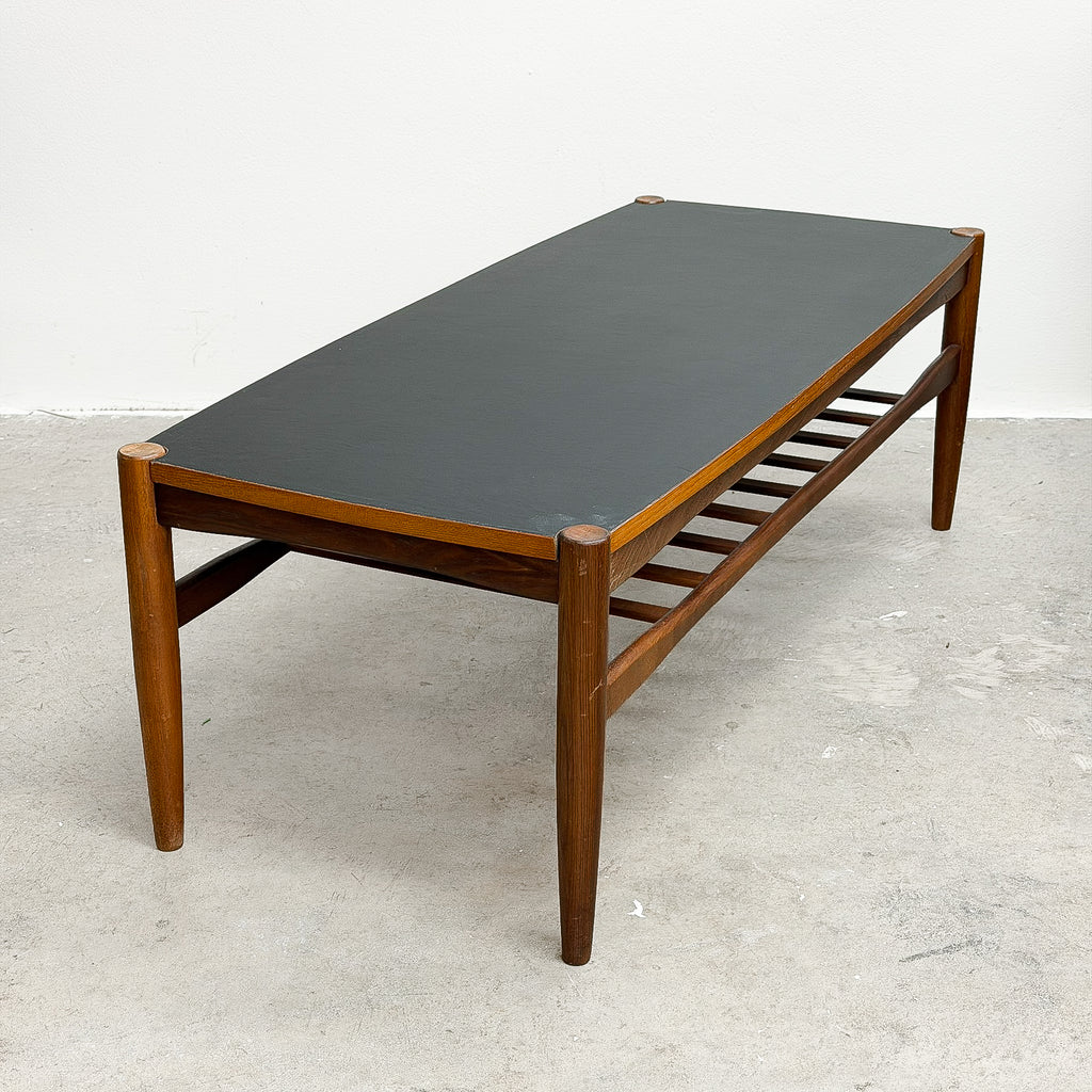 A mid-century modern wooden coffee table with a dark gray rectangular top and a lower slatted shelf, set against a plain white wall and concrete floor.