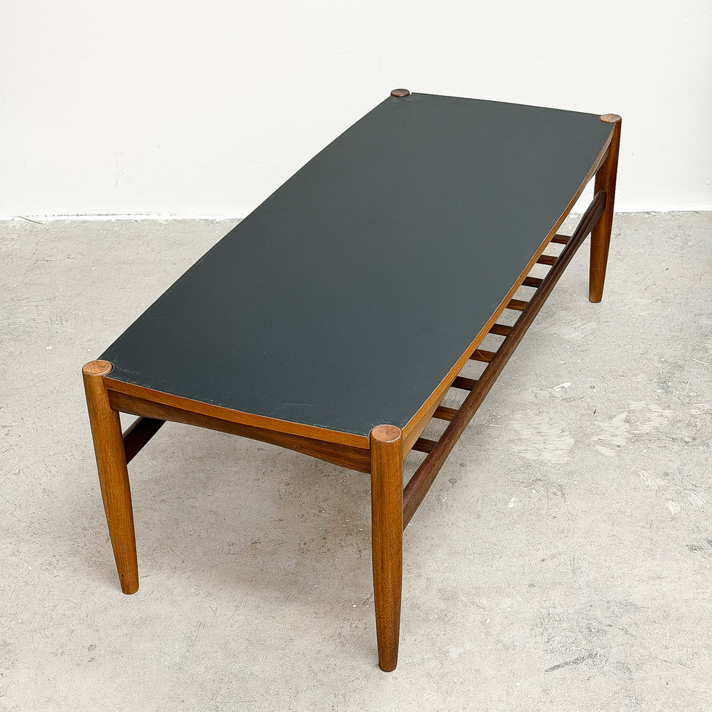 A mid-century modern wooden bench with a dark charcoal gray leather top and open slatted sides, photographed in a minimalist studio setting with a concrete floor and white wall.