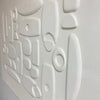 White textured wall art featuring a 3D abstract relief pattern of organic shapes and circles on a painted plaster-like surface.
