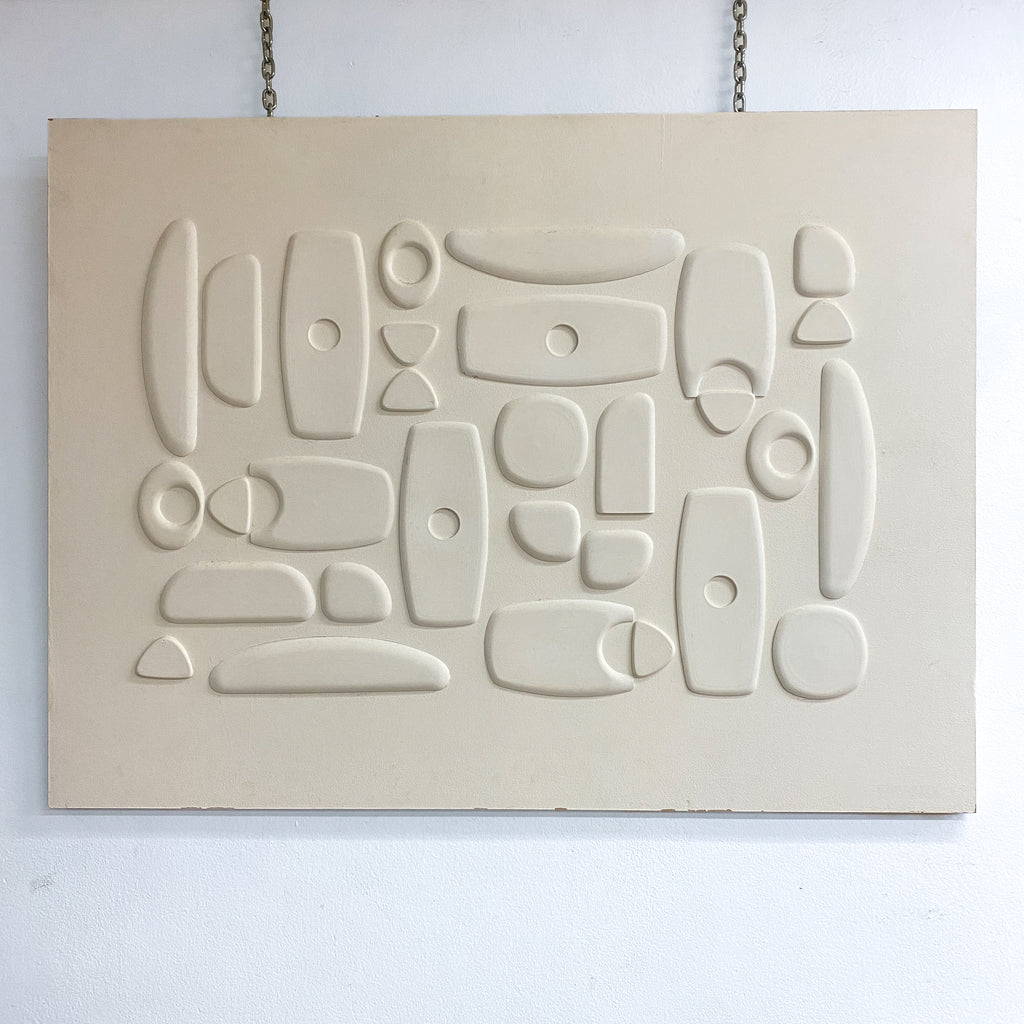 An abstract beige relief wall art panel with organic geometric shapes, hanging from two metal chains against a white wall.