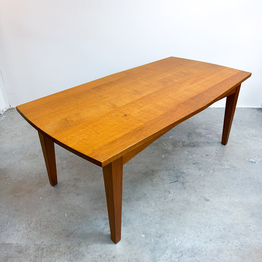 A mid-century modern wooden dining table with a warm honey-brown finish, featuring a gently curved apron and tapered legs, photographed in a warehouse setting.