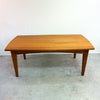 A mid-century modern wooden dining table with a warm honey finish, shown against a plain white wall and concrete floor.