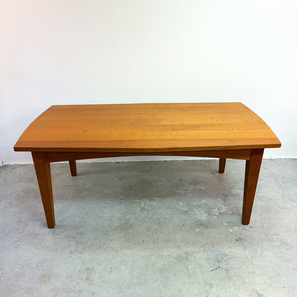 A mid-century modern wooden dining table with a warm honey finish, shown against a plain white wall and concrete floor.