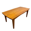 A mid-century modern wooden dining table with a warm honey-brown finish, featuring a gently curved apron and tapered legs, photographed in a warehouse setting.