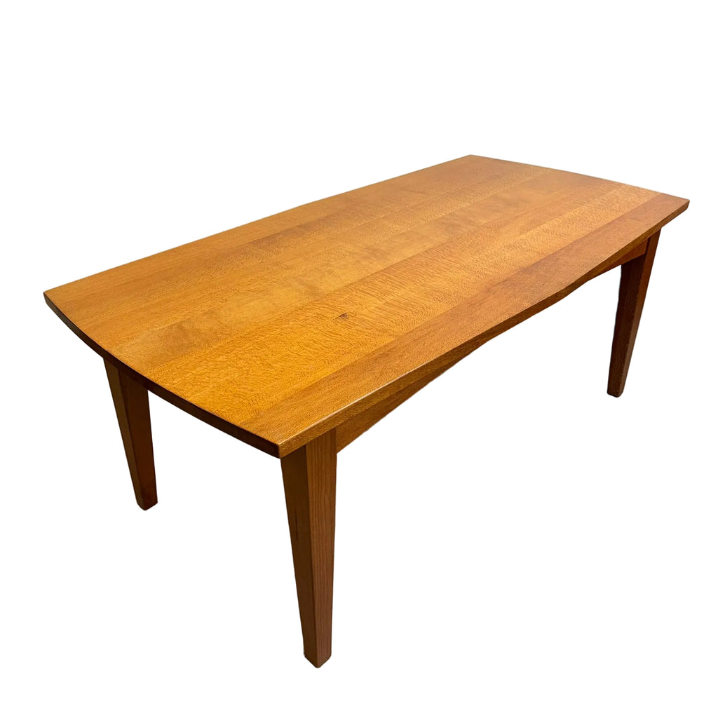 A mid-century modern wooden dining table with a warm honey-brown finish, featuring a gently curved apron and tapered legs, photographed in a warehouse setting.