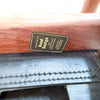Close-up of a wooden furniture leg showing a black metal brand plaque that reads 'Selected as Good Design for Australian Outdoor Furniture' and 'Industrial Strength of Australia', above a black leather runner with double-stitched detailing.