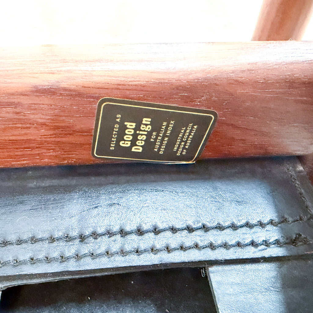 Close-up of a wooden furniture leg showing a black metal brand plaque that reads 'Selected as Good Design for Australian Outdoor Furniture' and 'Industrial Strength of Australia', above a black leather runner with double-stitched detailing.