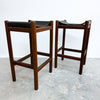 Two mid-century modern wooden bar stools with black leather seats, shown on a concrete floor against a white wall.