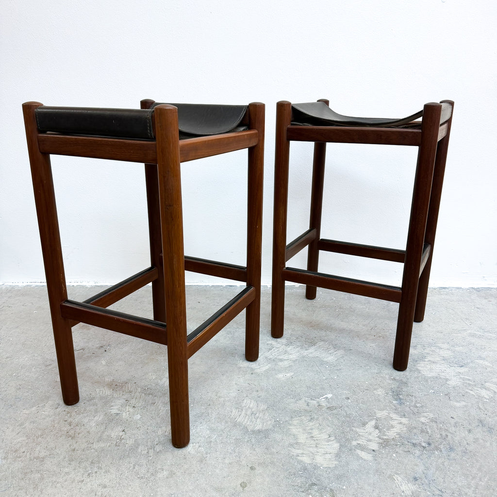 Two mid-century modern wooden bar stools with black leather seats, shown on a concrete floor against a white wall.