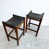 Two mid-century modern wooden bar stools with black leather seats, shown on a concrete floor against a white wall.
