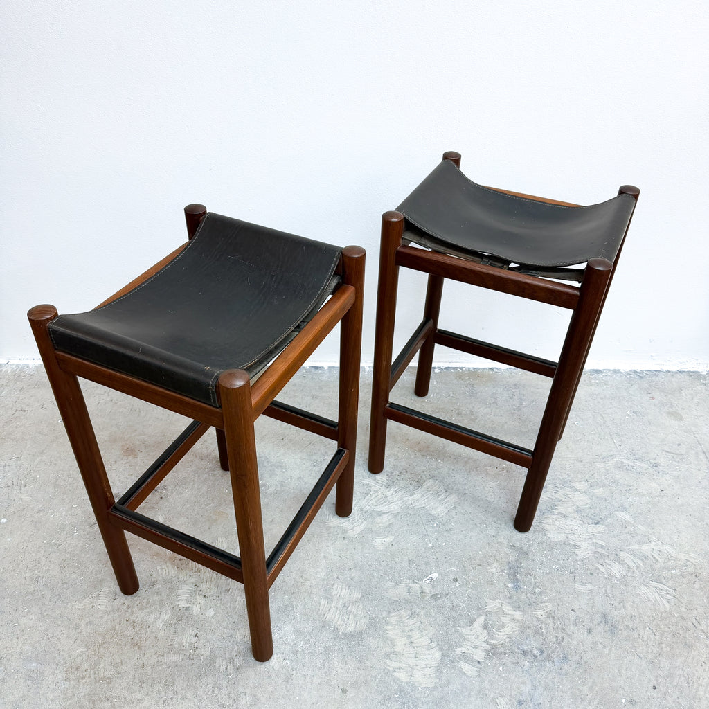 Two mid-century modern wooden bar stools with black leather seats, shown on a concrete floor against a white wall.