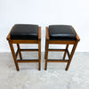 Two wooden bar stools with black cushioned seats, shown side by side against a plain white wall and concrete floor.