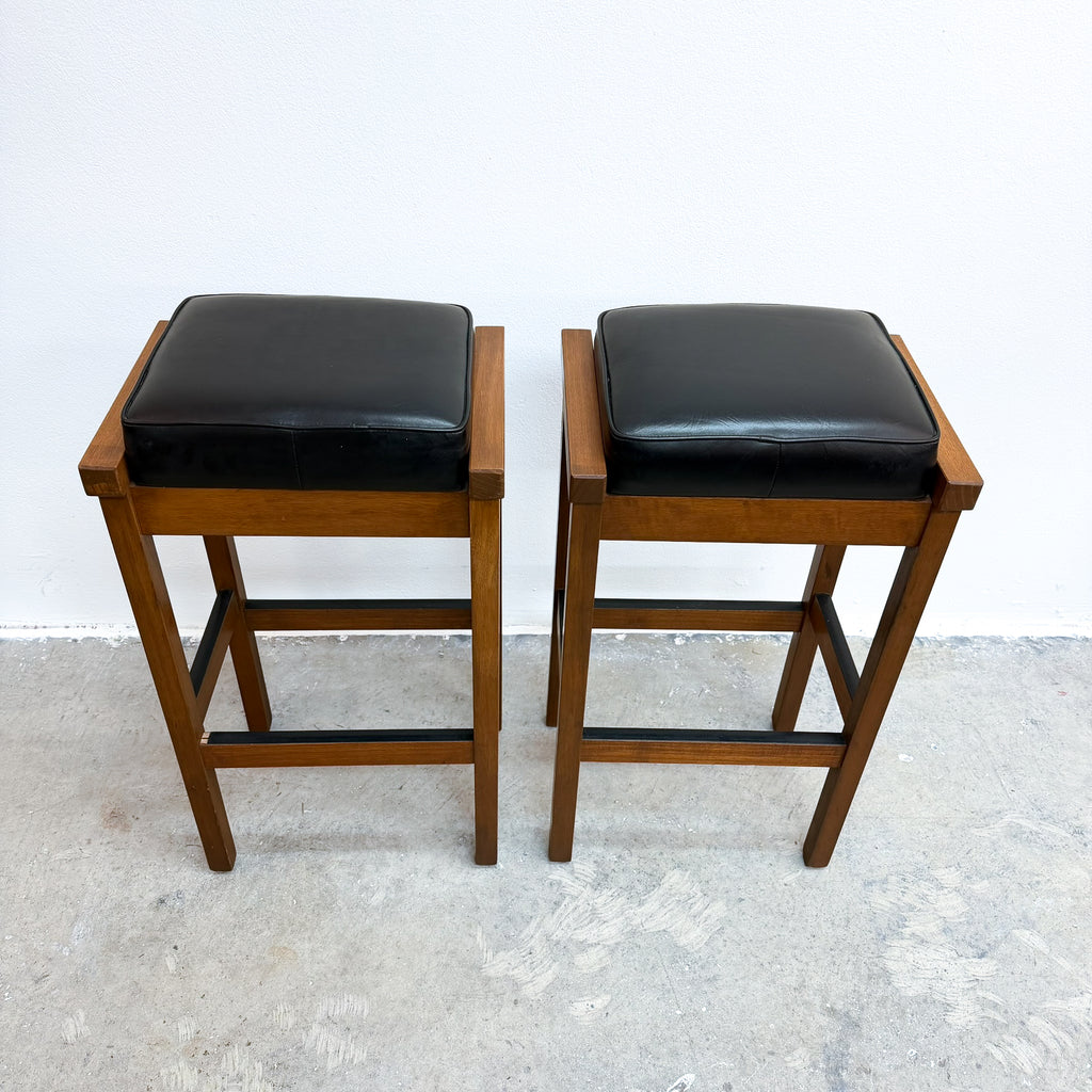 Two wooden bar stools with black cushioned seats, shown side by side against a plain white wall and concrete floor.