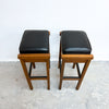 Two wooden bar stools with black cushioned seats, shown side by side against a plain white wall and concrete floor.