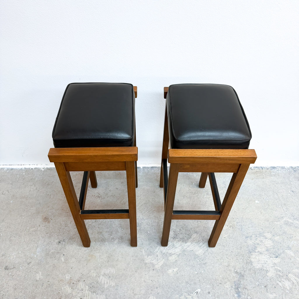 Two wooden bar stools with black cushioned seats, shown side by side against a plain white wall and concrete floor.