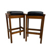 Two wooden bar stools with black cushioned seats, shown side by side against a plain white wall and concrete floor.