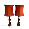 A pair of vintage table lamps with orange-red fabric drum shades and dark brown turned wood bases, standing on a concrete floor against a white wall.