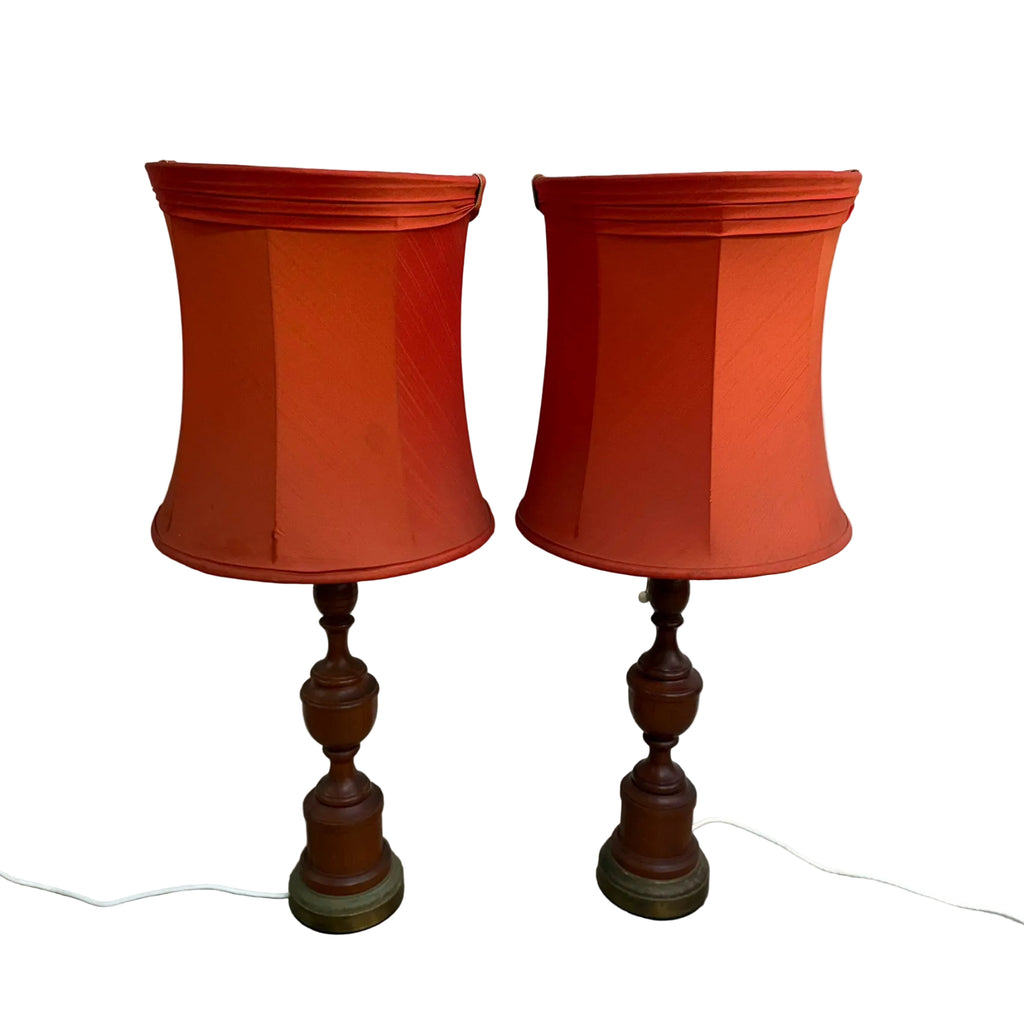 A pair of vintage table lamps with orange-red fabric drum shades and dark brown turned wood bases, standing on a concrete floor against a white wall.