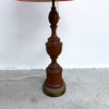 A vintage-style wooden table lamp with a multi-tiered turned wood base in a warm brown finish, standing on a concrete floor against a white wall.