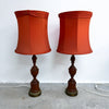 A pair of vintage table lamps with rust-orange fabric drum shades and dark brown turned wood bases, standing on a concrete floor against a white wall.