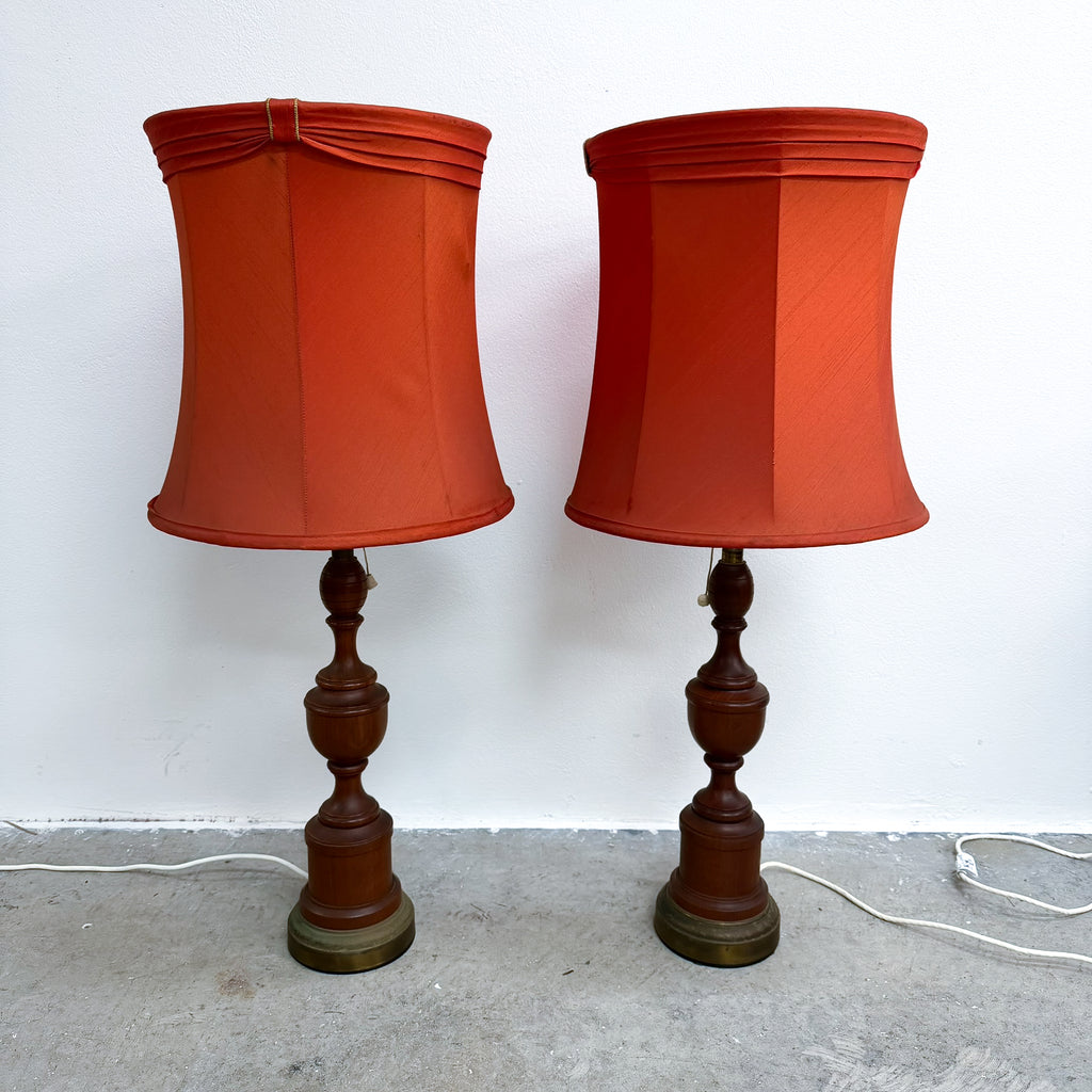 A pair of vintage table lamps with rust-orange fabric drum shades and dark brown turned wood bases, standing on a concrete floor against a white wall.