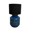 A table lamp with a black fabric drum shade and a glossy cobalt blue ceramic base decorated with gold geometric patterns, set against a white wall and gray floor.