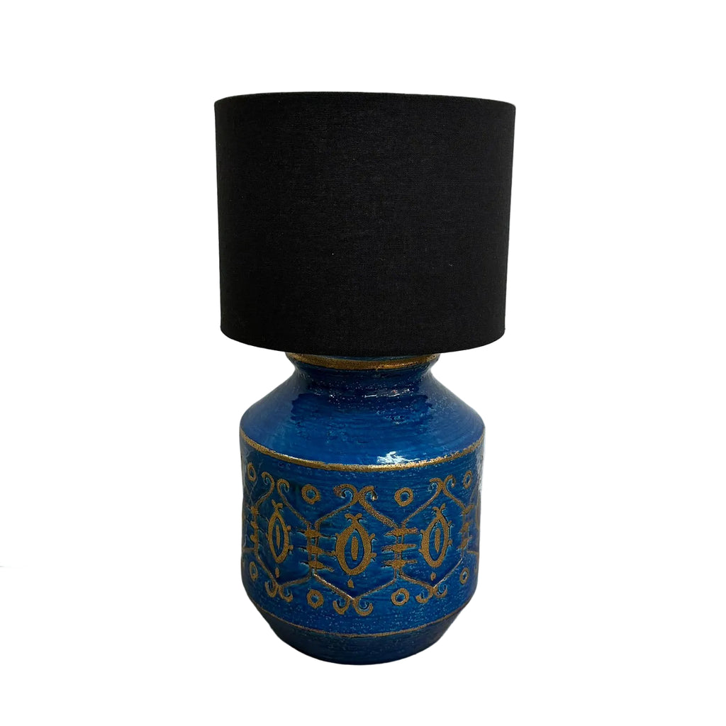 A table lamp with a black fabric drum shade and a glossy cobalt blue ceramic base decorated with gold geometric patterns, set against a white wall and gray floor.
