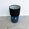A table lamp with a black fabric drum shade resting on a glossy blue ceramic base decorated with gold geometric patterns, set against a white wall and concrete floor.
