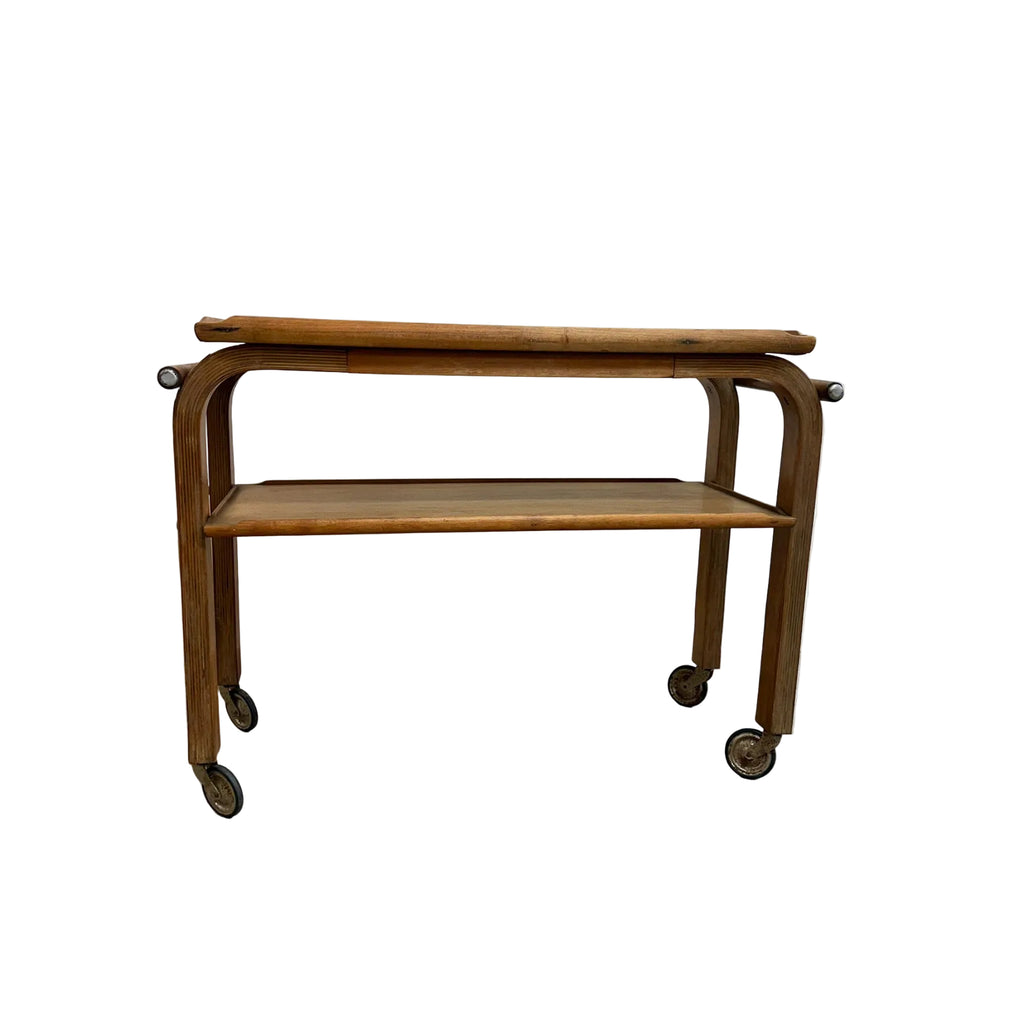 A vintage wooden serving cart with two shelves and caster wheels, photographed against a plain white wall and gray concrete floor.