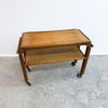 A mid-century modern wooden serving cart with a warm medium-brown finish, featuring a top tray, a lower shelf, and dark metal caster wheels, set against a plain white wall and gray concrete floor.
