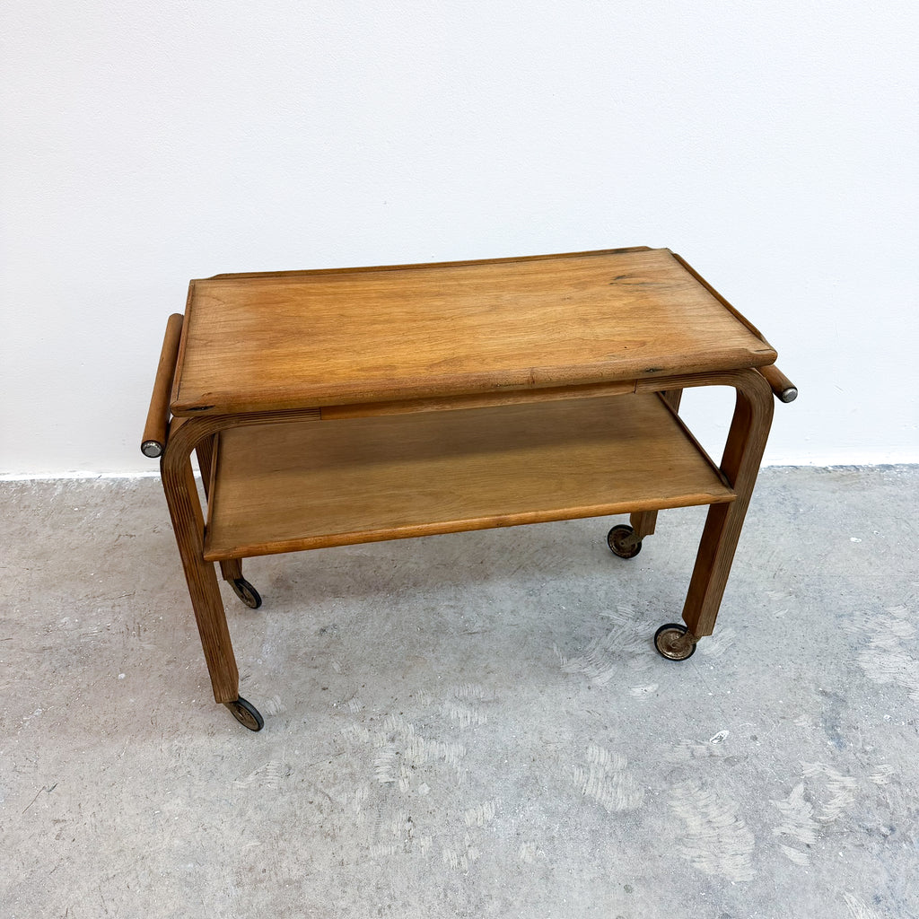 A mid-century modern wooden serving cart with a warm medium-brown finish, featuring a top tray, a lower shelf, and dark metal caster wheels, set against a plain white wall and gray concrete floor.
