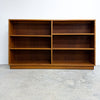 A mid-century modern wooden bookcase with a warm medium-brown finish, featuring six open shelves arranged in two columns of three, set against a plain white wall and concrete floor.