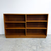 A mid-century modern wooden bookcase with a warm medium-brown finish, featuring six open shelves arranged in two columns of three, set against a plain white wall and concrete floor.