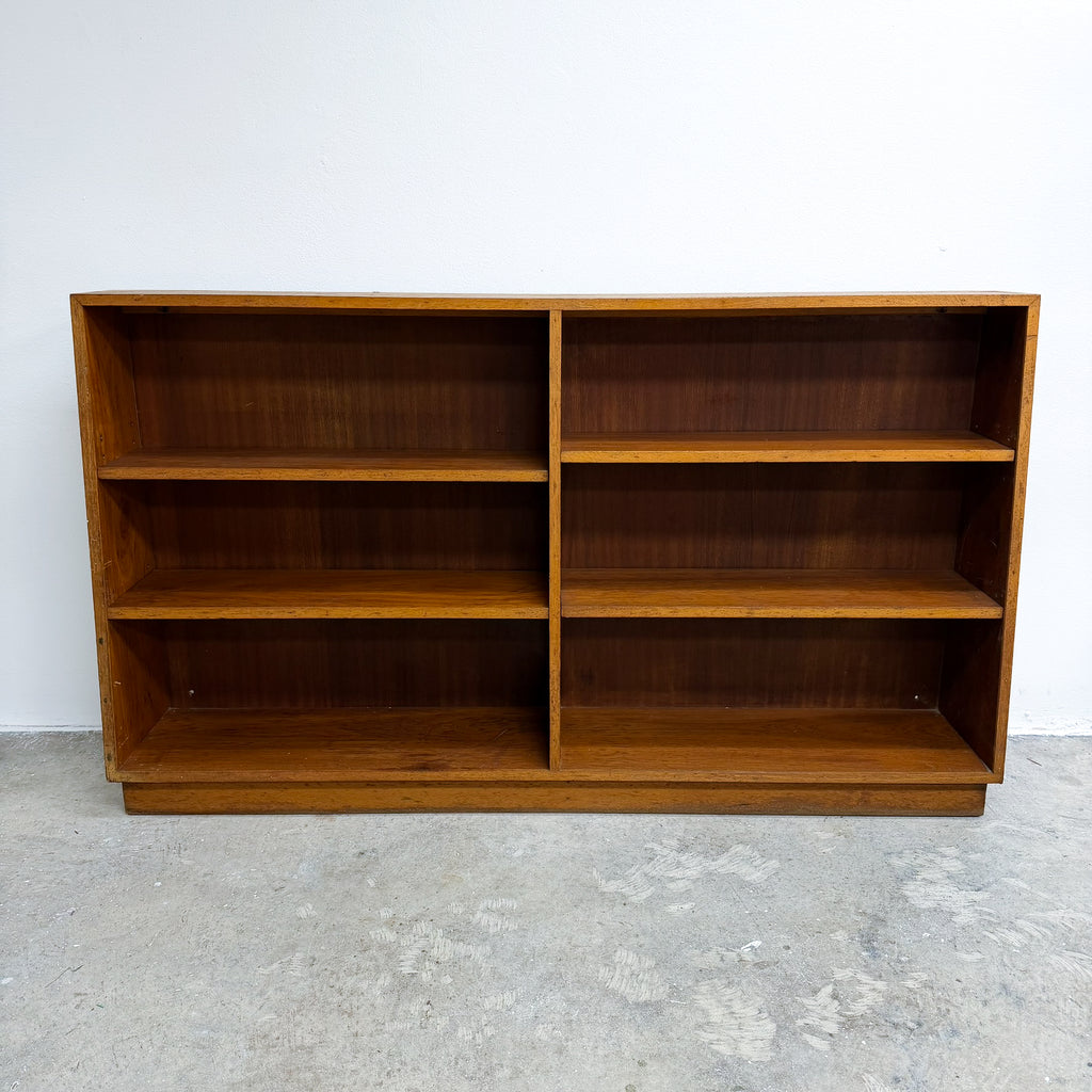 A mid-century modern wooden bookcase with a warm medium-brown finish, featuring six open shelves arranged in two columns of three, set against a plain white wall and concrete floor.