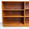 A wooden bookcase with multiple open shelves, shown from the front against a plain white wall and light gray floor.