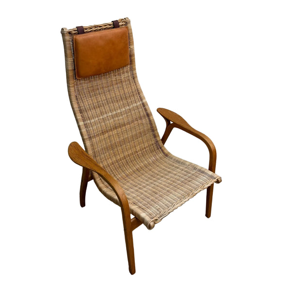 A mid-century modern lounge chair with a woven rattan seat and back, curved wooden arms, and a brown leather headrest, shown on a concrete floor against a white wall.