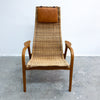 A mid-century modern lounge chair with a woven wicker seat and backrest, featuring a brown leather headrest and curved wooden arms and legs, set against a plain white wall and gray concrete floor.