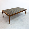 A mid-century modern rectangular coffee table with a dark mosaic tile top and a warm brown wood frame, photographed in a minimalist studio setting.