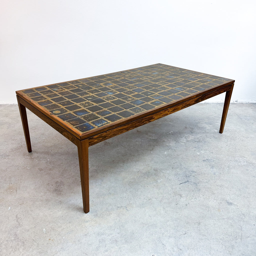 A mid-century modern rectangular coffee table with a dark mosaic tile top and a warm brown wood frame, photographed in a minimalist studio setting.