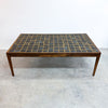 A mid-century modern coffee table with a rectangular wooden frame and a top composed of small dark blue-gray tiles with gold grid lines, set against a plain white wall and concrete floor.