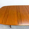 A close-up of a mid-century modern wooden dining table with a warm honey-brown grain finish and a visible extension leaf on the right side, set against a plain white wall and concrete floor.