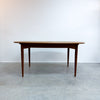 A mid-century modern wooden dining table with a warm brown finish, shown against a plain white wall and gray concrete floor.