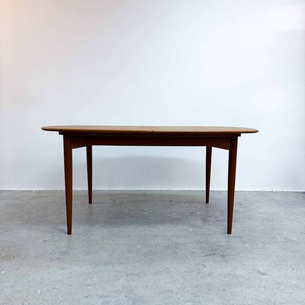 A mid-century modern wooden dining table with a warm brown finish, shown against a plain white wall and gray concrete floor.