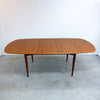A mid-century modern wooden dining table with rounded ends and tapered legs, shown on a concrete floor against a plain white wall.