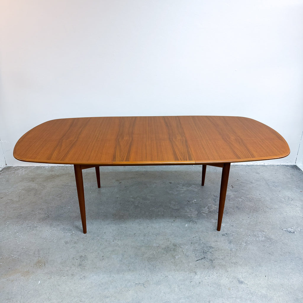 A mid-century modern wooden dining table with rounded ends and tapered legs, shown on a concrete floor against a plain white wall.