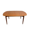 A mid-century modern wooden dining table with a warm medium-brown wood grain finish, featuring a rounded rectangular top and tapered legs, set against a plain white wall and concrete floor.