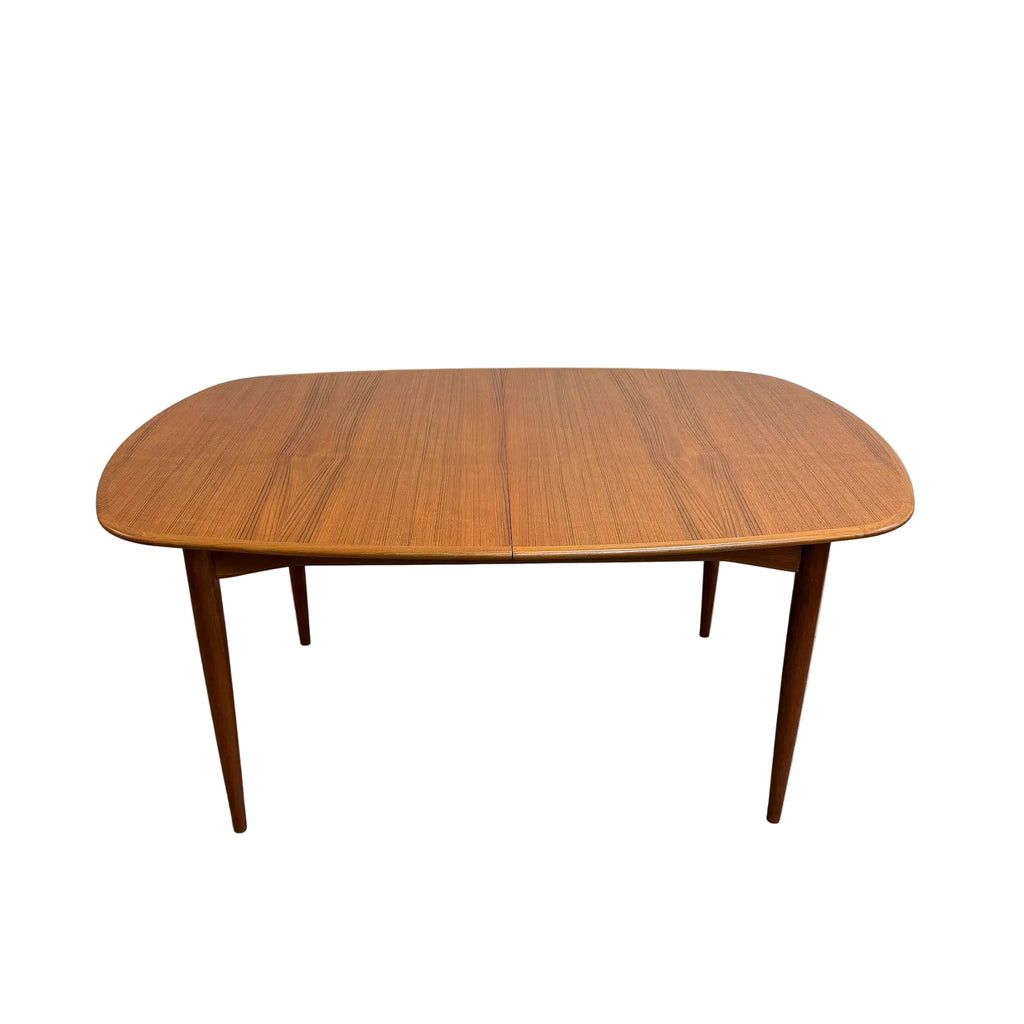 A mid-century modern wooden dining table with a warm medium-brown wood grain finish, featuring a rounded rectangular top and tapered legs, set against a plain white wall and concrete floor.