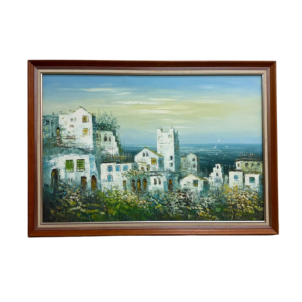 A framed painting of a coastal village with white buildings, blue sky, and sea, displayed against a white wall on a gray concrete floor.