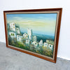 A framed landscape painting of a coastal town with white buildings, greenery, and a pale blue sky, shown leaning against a white wall on a gray concrete floor.